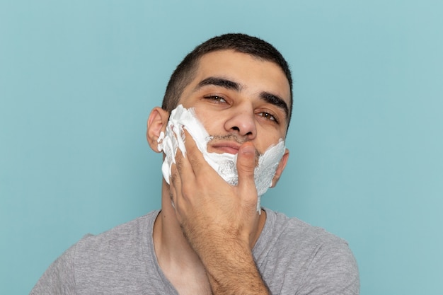 Vue rapprochée avant jeune homme en t-shirt gris couvrant son visage avec de la mousse blanche pour le rasage sur le rasoir en mousse de barbe bureau bleu glace