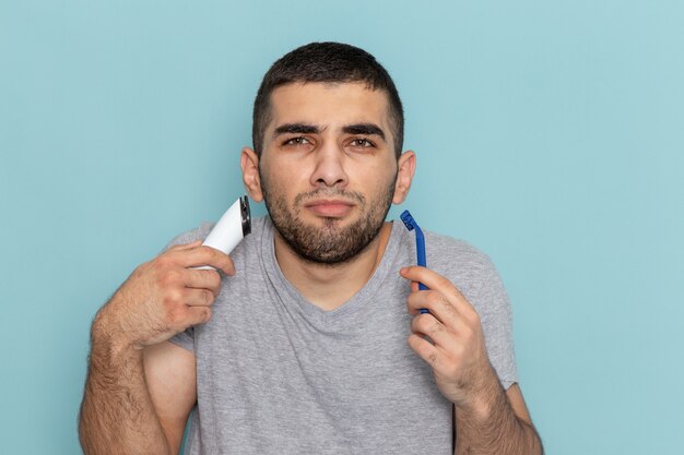 Vue rapprochée avant jeune homme en t-shirt gris tenant le rasoir et le rasoir électrique sur le mur bleu rasage barbe cheveux couleur mousse