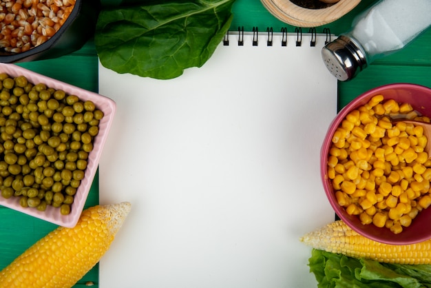 Vue rapprochée de bols de graines de maïs et de pois verts avec du sel d'épinards cors et bloc-notes sur fond vert avec copie espace