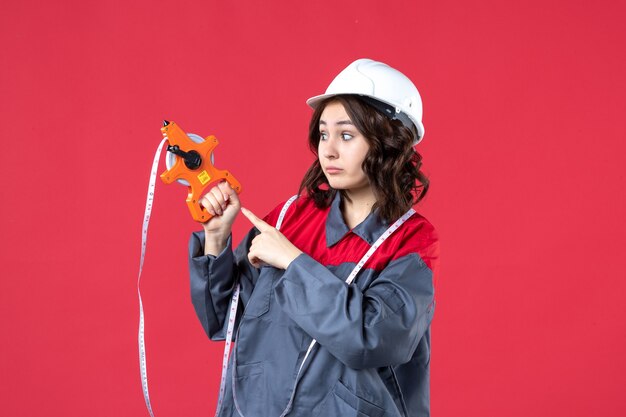 Vue rapprochée de curieuse femme architecte en uniforme portant un casque avec ruban à mesurer sur mur rouge