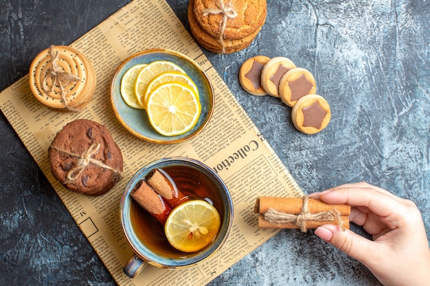 Vue rapprochée de délicieux biscuits et main tenant une tasse de thé noir à la cannelle sur un vieux journal sur fond sombre