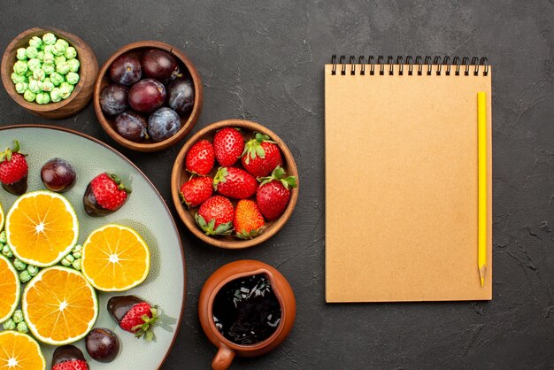 Vue rapprochée de dessus fruits bonbons verts fraise enrobée de chocolat orange hachée et bols de baies et sauce au chocolat à côté du cahier et du crayon