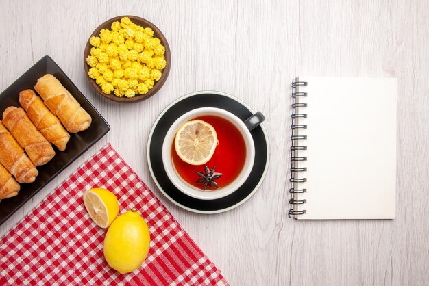 Vue rapprochée de dessus la pâtisserie sur l'assiette assiette de pâtisserie à côté du citron sur la nappe à carreaux blanc bol de cahier de bonbons jaunes et une tasse de thé sur la table blanche