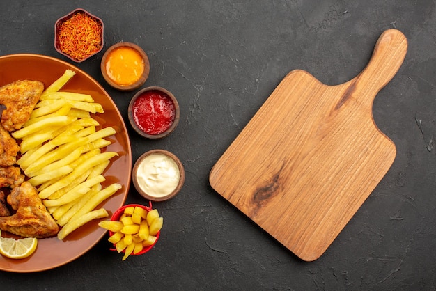 Vue rapprochée de dessus poulet et pommes de terre ailes de poulet frites et citron trois bols de différents types de sauces et d'épices à côté de la planche à découper en bois sur la table sombre