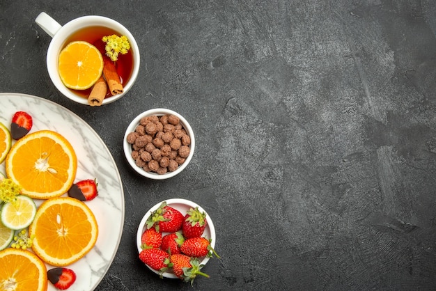 Vue rapprochée de dessus une tasse de thé et de fruits une tasse de thé avec des bâtons de citron et de cannelle des bols de noix et de fraises et une assiette d'agrumes et de fraises enrobées de chocolat