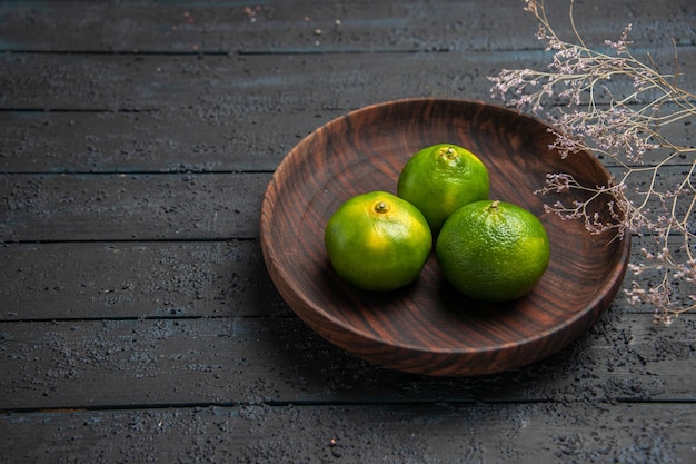 Vue rapprochée de dessus trois citrons verts dans un bol trois citrons verts dans un bol marron à côté des branches sur la table sombre