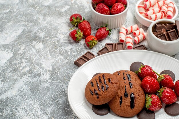 Vue rapprochée du bas des cookies fraises et chocolats ronds sur la plaque blanche et bols de bonbons chocolats fraises sur la table gris-blanc