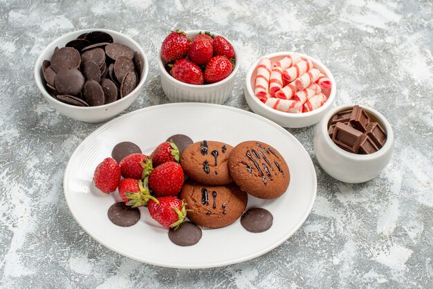 Vue rapprochée du bas des cookies fraises et chocolats ronds sur la plaque ovale blanche entourée de bols avec des bonbons fraises et chocolats sur le terrain avec copie espace