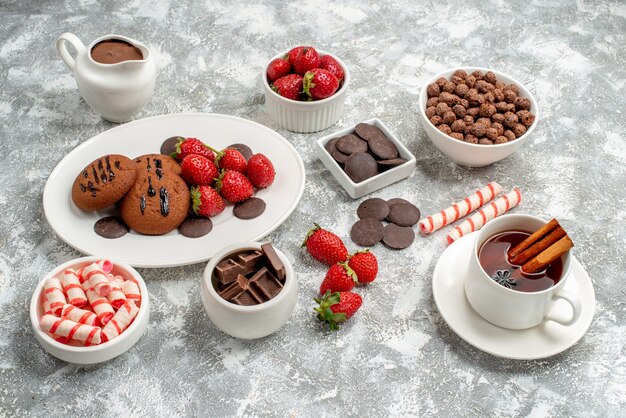 Vue rapprochée du bas cookies fraises et chocolats ronds sur la plaque ovale bols de bonbons fraises chocolats céréales cacao et cannelle thé aux graines d'anis sur le tableau gris-blanc