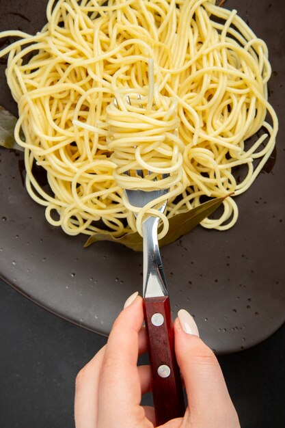 Vue rapprochée du bas des pâtes spaghetti aux feuilles de laurier sur une fourchette à assiette en main féminine sur fond noir