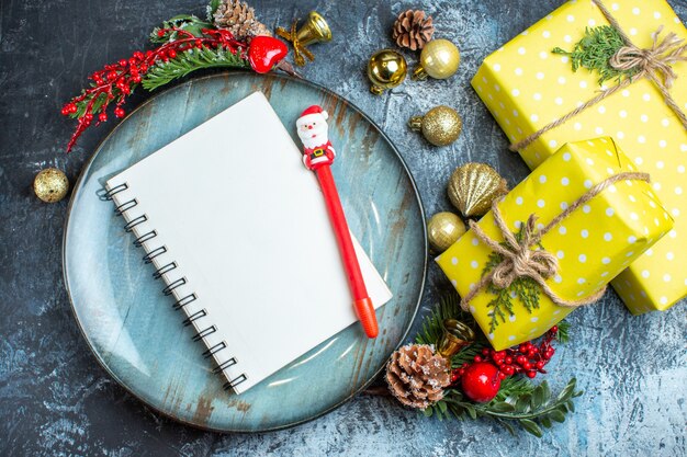 Vue rapprochée du cahier avec stylo et coffrets cadeaux sur une plaque bleue accessoires décoratifs cône de conifères à côté de coffrets cadeaux jaunes sur fond sombre