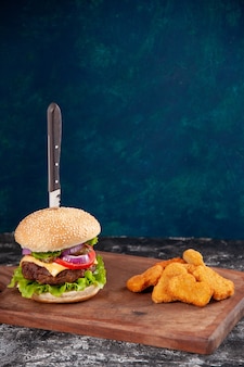 Vue rapprochée du couteau dans un délicieux sandwich à la viande et des pépites de poulet sur une planche de bois sur une surface bleu foncé