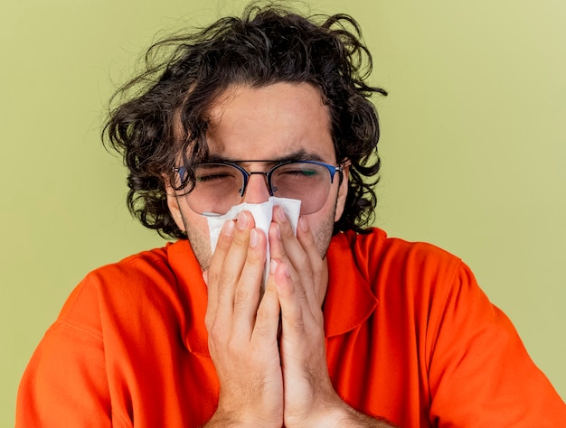 Vue rapprochée du jeune homme malade portant des lunettes essuyant le nez avec serviette avec les yeux fermés isolé sur mur vert olive