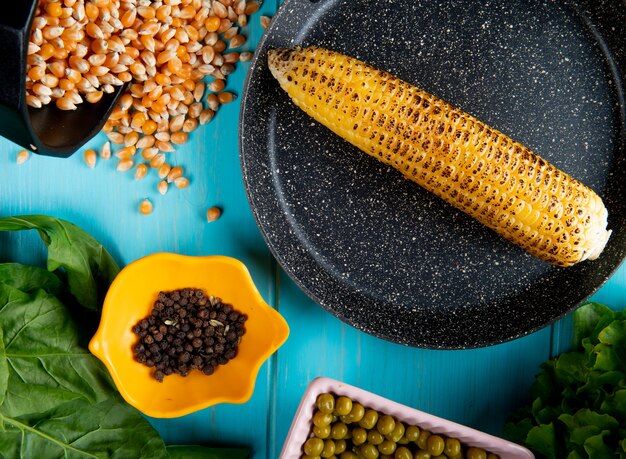 Vue rapprochée d'épis de maïs dans une casserole avec des graines de maïs, des graines de poivre noir et de la laitue sur la surface bleue