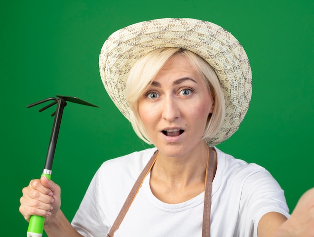 Vue rapprochée d'une femme jardinière blonde d'âge moyen en uniforme portant un chapeau tenant un râteau houe tendant la main isolée sur un mur vert