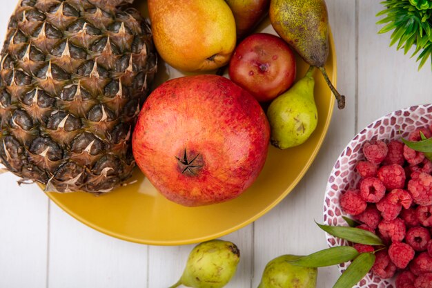 Vue rapprochée de fruits comme la pêche à la grenade ananas dans la plaque avec des framboises dans un bol sur la surface en bois