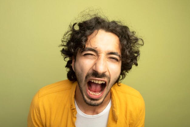 Vue rapprochée de furieux jeune bel homme caucasien regardant la caméra crier isolé sur fond vert olive