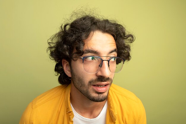 Vue rapprochée de impressionné jeune bel homme caucasien portant des lunettes à côté isolé sur mur vert olive