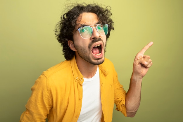 Photo gratuite vue rapprochée de impressionné jeune bel homme caucasien portant des lunettes pointant sur le côté isolé sur mur vert olive