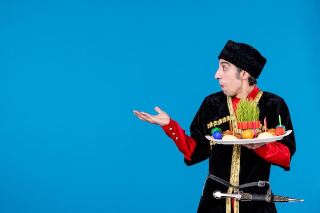 Vue rapprochée de l'interrogatoire d'un jeune homme en costume traditionnel tenant un plateau rempli de confiseries nationales et pointant quelque chose sur le côté droit sur fond bleu