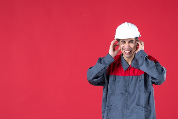 Vue rapprochée d'un jeune architecte nerveux en uniforme avec un casque sur un mur rouge isolé