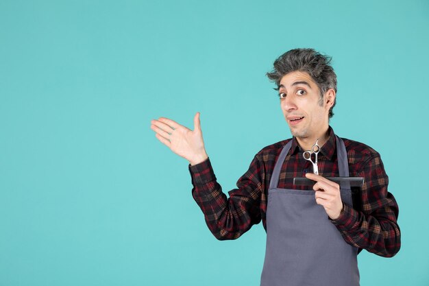 Vue rapprochée d'un jeune coiffeur curieux portant un tablier gris et tenant un peigne et des ciseaux pointant quelque chose sur le côté droit sur fond bleu