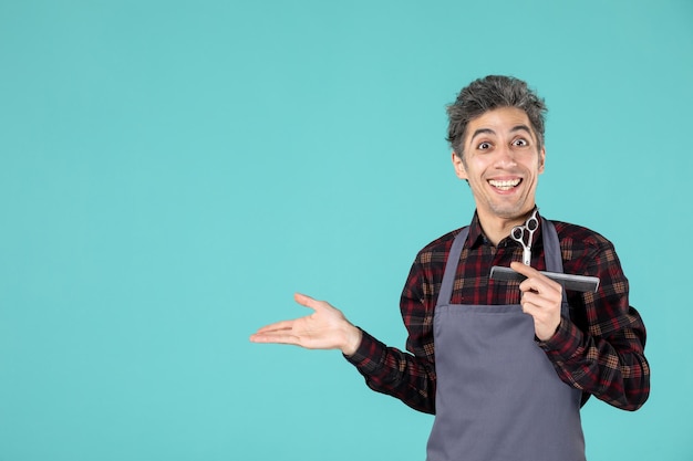 Vue Rapprochée D'un Jeune Coiffeur Souriant Portant Un Tablier Gris Et Tenant Un Peigne Et Des Ciseaux Pointant Quelque Chose Sur Le Côté Droit Sur Fond Bleu