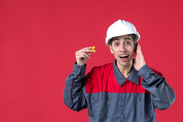 Vue rapprochée d'un jeune constructeur concentré en uniforme avec un casque et tenant des bouchons d'oreille sur un mur rouge isolé