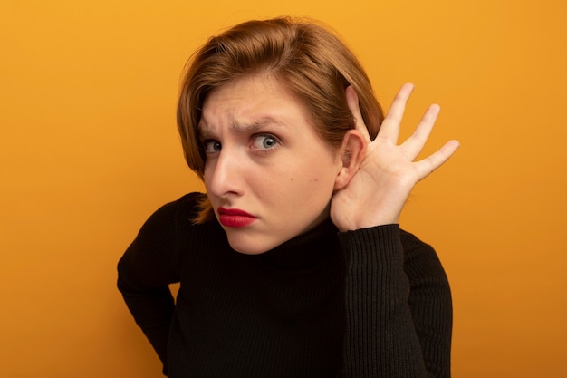 Vue rapprochée d'une jeune fille blonde curieuse gardant la main sur la taille mettant une autre main derrière l'oreille faisant je ne peux pas t'entendre geste isolé sur le mur orange