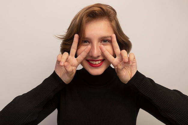 Photo gratuite vue rapprochée d'une jeune fille blonde souriante gardant les mains devant le visage faisant un signe de paix isolé sur un mur blanc