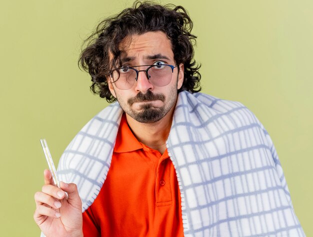 Vue rapprochée d'un jeune homme malade anxieux portant des lunettes enveloppées dans un thermomètre à carreaux holding à l'avant isolé sur mur vert olive