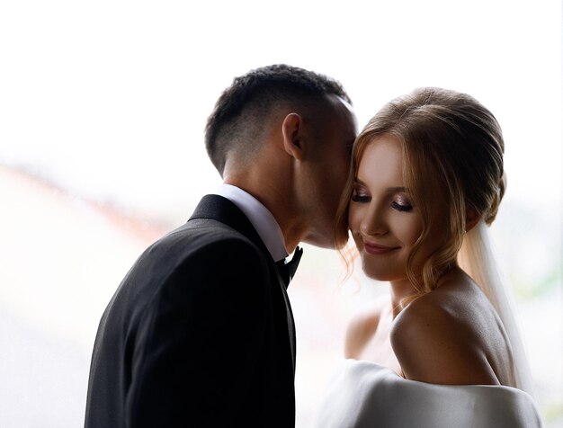 Vue rapprochée d'une jolie femme avec une coiffure élégante vêtue d'une robe blanche avec des épaules nues souriant et regardant ailleurs tandis que le marié sans visage embrassant son visage pendant la célébration du mariage