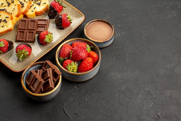 Vue rapprochée latérale dessert appétissant gâteau aux fraises et chocolat sur plaque grise à côté de bols avec fraises et crème au chocolat sur le côté gauche de la table