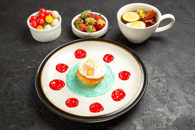 Vue rapprochée latérale du cupcake et du cupcake aux fraises avec de la crème blanche et des sauces colorées à côté des bonbons et des fraises dans des bols et une tasse de tisane sur la table sombre