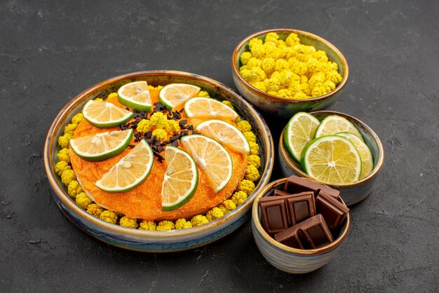 Vue rapprochée latérale du gâteau aux agrumes gâteau appétissant aux agrumes et au chocolat à côté des bols de citrons verts tranchés au chocolat et de bonbons jaunes sur le mur sombre