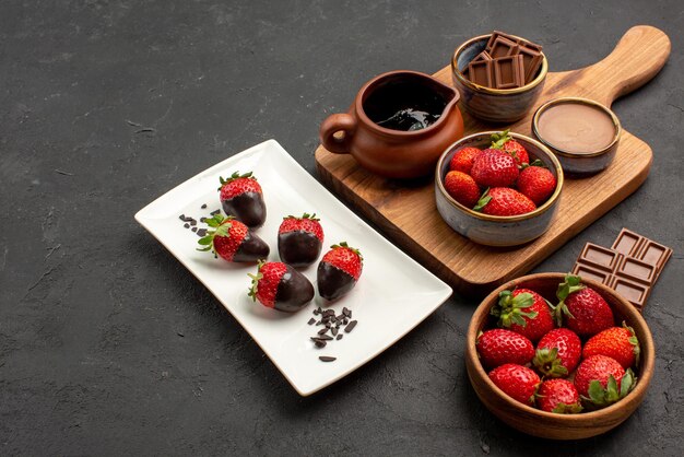 Vue rapprochée latérale fraises barres appétissantes de chocolat bol de fraises assiette de fraises enrobées de chocolat et planche de cuisine avec crème au chocolat et fraises