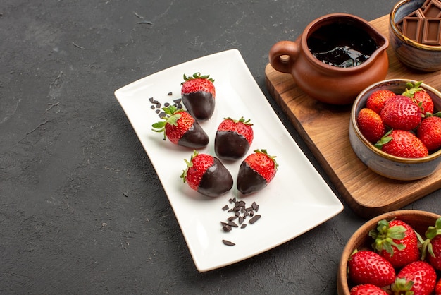 Vue rapprochée latérale des fraises enrobées de chocolat assiette de fraises enrobées de chocolat barres de chocolat bol de fraises et planche de cuisine avec crème au chocolat et fraises