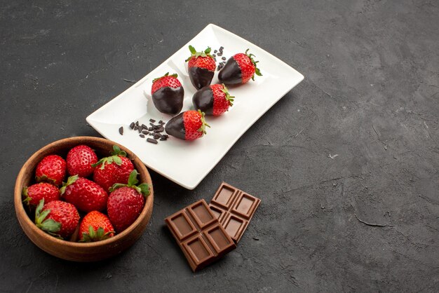 Vue rapprochée latérale des fraises enrobées de chocolat bol de fraises et barres de chocolat à côté de l'assiette de fraises enrobées de chocolat sur la table sombre