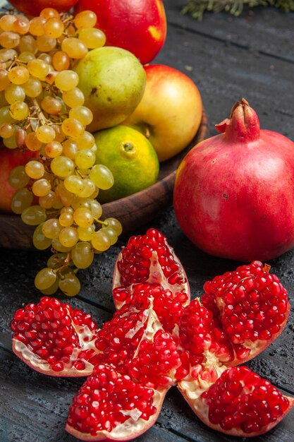 Vue rapprochée latérale des fruits dans une assiette brune assiette de raisins blancs et noirs limes poires pommes à côté de grenade et de branches pilées sur une table grise