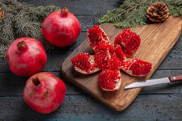 Vue rapprochée latérale des grenades rouges pilées de grenade sur une planche de cuisine à côté de branches avec un couteau à cônes et trois grenades sur une table grise