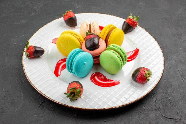 Vue rapprochée latérale des macarons une assiette de macarons colorés, de fraises enrobées de chocolat et de sauce sur la table sombre