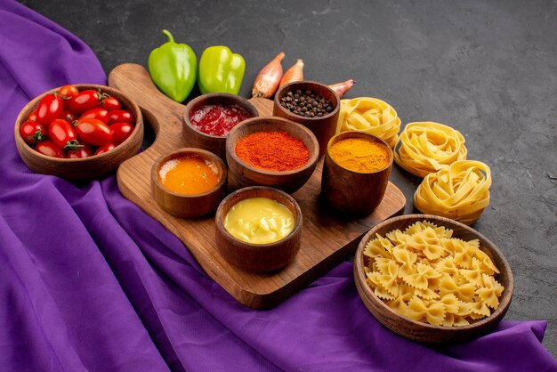 Vue rapprochée latérale des pâtes et des épices, des oignons verts et des bols de tomates à côté des épices et des sauces sur la planche de bois et des pâtes sur la nappe violette