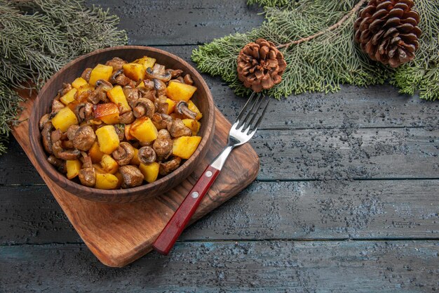 Vue rapprochée latérale plat et planche à découper bol marron de champignons et pommes de terre à côté d'une fourchette et d'une planche à découper sous des branches d'épinette avec des cônes