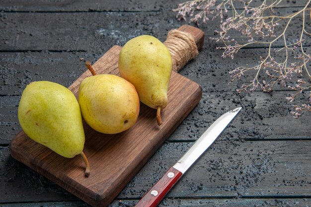 Vue rapprochée latérale des poires à bord de trois poires vert-jaune-rouge sur une planche à découper au centre d'une table sombre à côté d'un couteau et de branches d'arbre