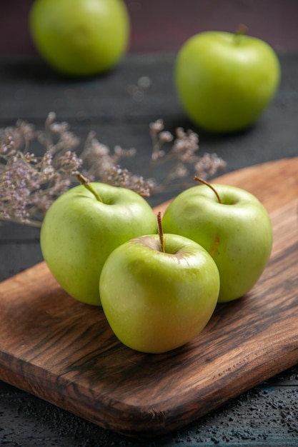 Vue rapprochée latérale des pommes à bord de trois pommes vertes sur une planche de cuisine à côté de deux pommes et de branches d'arbres sur une table sombre