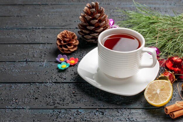 Vue rapprochée latérale d'une tasse de branches d'épinette à thé avec des jouets de Noël et des cônes à côté de la tasse de thé sur la soucoupe blanche, des bâtons de cannelle et du citron sur la table