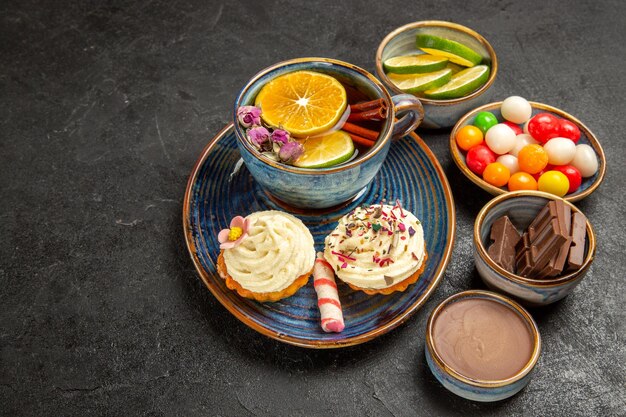 Vue rapprochée latérale tisane une tasse de thé au citron et cupcakes à la crème à côté des bols de tranches de chocolat de crème au chocolat au citron vert et de bonbons colorés sur la table