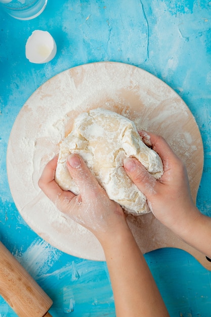 Vue rapprochée de la main de femme pétrir la pâte sur une planche à rouler avec coquille d'oeuf sur bleu