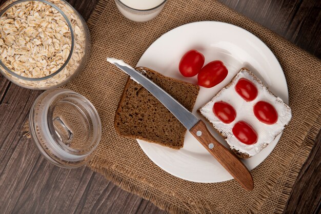 Vue rapprochée de la plaque avec des tranches de pain de seigle enduites de fromage cottage et de tomates et d'un couteau avec des flocons d'avoine sur fond de bois