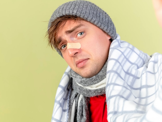 Vue rapprochée de triste jeune homme malade blond beau portant un chapeau d'hiver et une écharpe enveloppée de plaid à l'avant avec du plâtre sur le nez isolé sur un mur vert olive